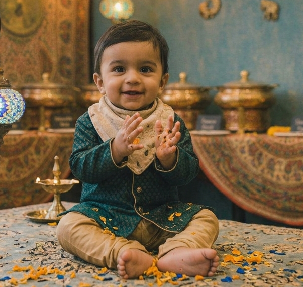 Baby Aveer sitting cross-legged in a teal embroidered kurta, clapping and smiling at the camera.